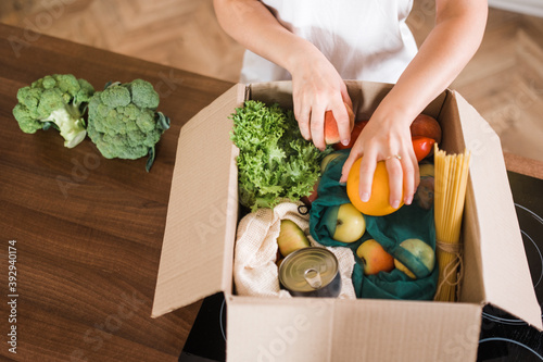 Home food delivery. Various vegetables, fruits and other products in a box that a young girl is sorting out at home on the kitchen table