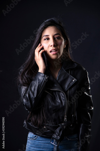 Young latin woman speaking through her cellphone in isolated in a dark background and wearing a black jacket