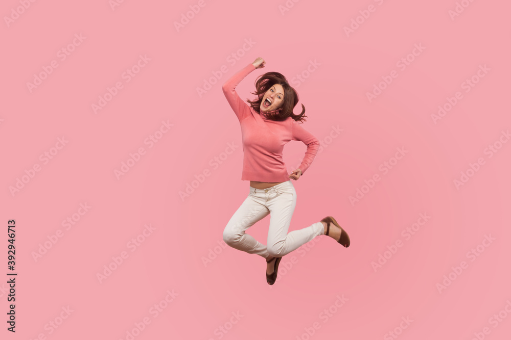 Extremely happy satisfied woman jumping high celebrating her success, flying in air with pleased smiling expression, dreams comes true. Indoor studio shot isolated on pink background
