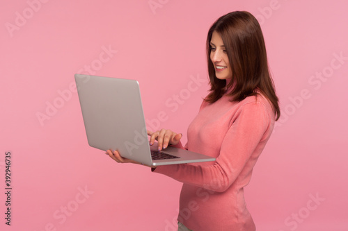 Smiling positive woman with brown hair in pink sweater working on laptop, freelance job, online studying. Indoor studio shot isolated on pink background