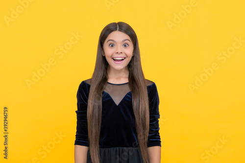 Suprised little kid with long hair in fashion dress keep mouth opened yellow background, surprise