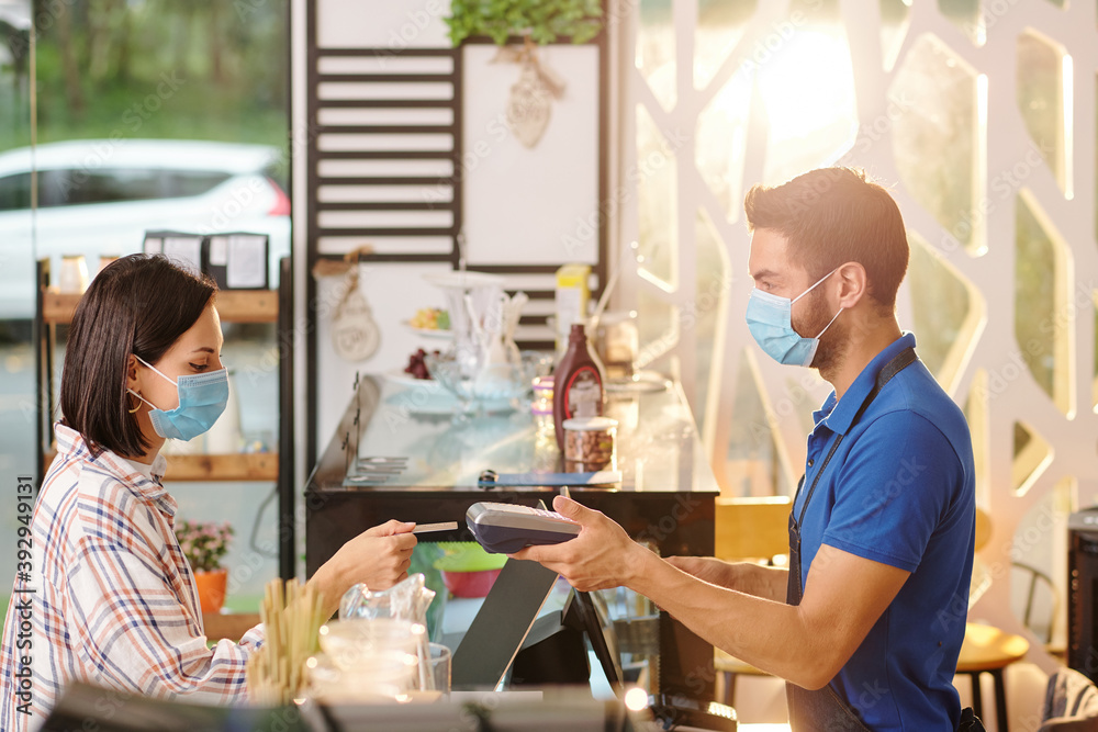 Pretty young woman in medical mask paying for order in cafe with credit card Pretty young woman in medical mask paying for order in cafe with credit card