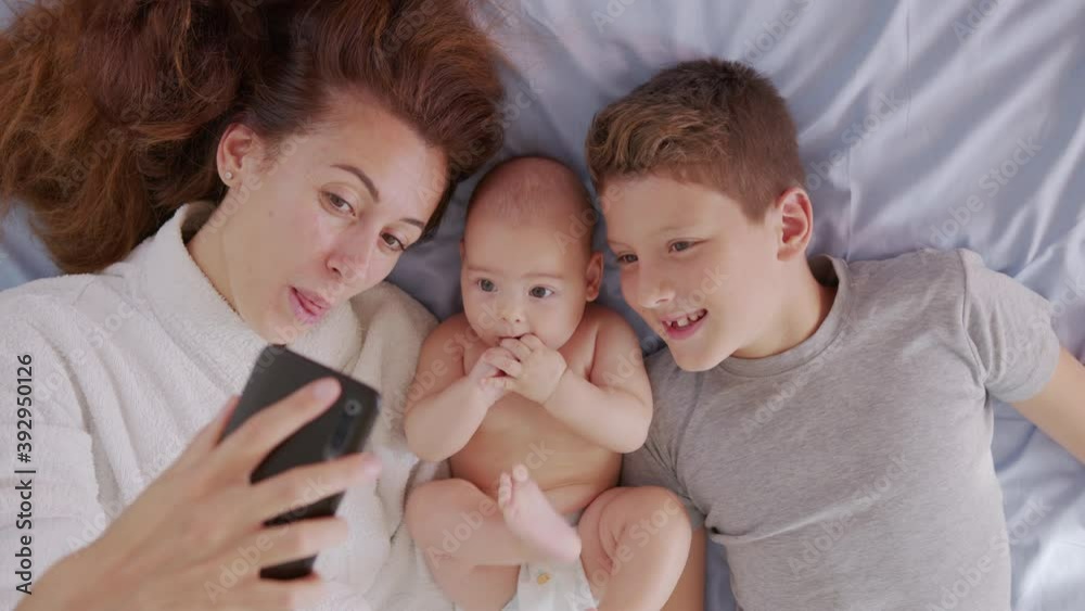 Happy mother with her kids are making a selfie or video call to father or relatives in a bed. Concept of technology, new generation,family, connection, parenthood, authenticity. Happy mother with her kids are making a selfie or video call to father or relatives in a bed. Concept of technology, new generation,family, connection, parenthood, authenticity.