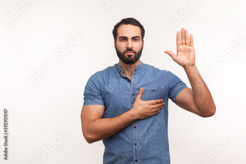 I swear! Conscious honest man raising one hand and holding on chest another, swearing in honesty and devotion, patriotism. Indoor studio shot isolated on white background photo