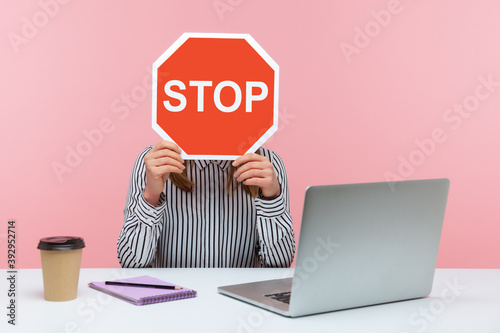 Foto Female office worker in striped shirt sitting at workplace hiding face behind red stop traffic sign avoiding conflicts, afraid of workplace bullying