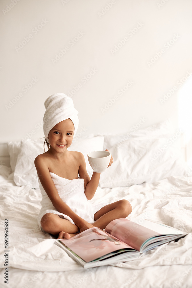 Little girl wrapped in a bath towel with a cup of milk in her hand reading a magazine. Little girl wrapped in a bath towel with a cup of milk in her hand reading a magazine.