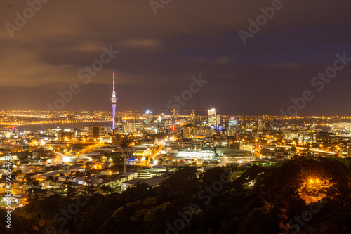 Auckland's illuminated skyline featuring the Sky Tower and modern high-rise buildings is captured at dusk, with harbour lights reflecting off the water.