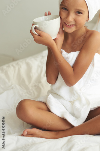 Beautiful little girl wrapped in bath towels drinks milk sitting on the bed.