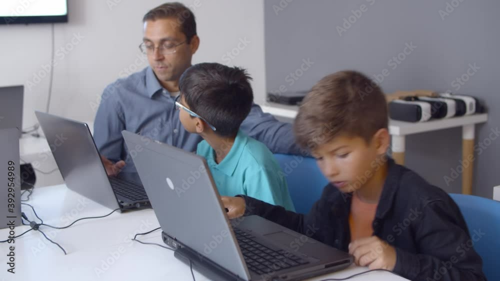 Computer science teacher teaching schoolboys to write codes, sitting at desk with children, helping and explaining task. Medium shot. Digital education concept Computer science teacher teaching schoolboys to write codes, sitting at desk with children, helping and explaining task. Medium shot. Digital education concept