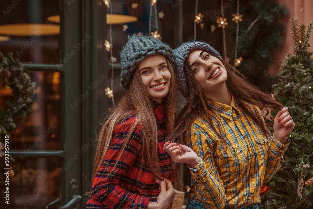 Christmas, New Year, winter holidays conception: two happy smiling women, friends, sisters, wearing knitted hat, tartan shirts, posing in street with festive decorations. Copy, empty space for text