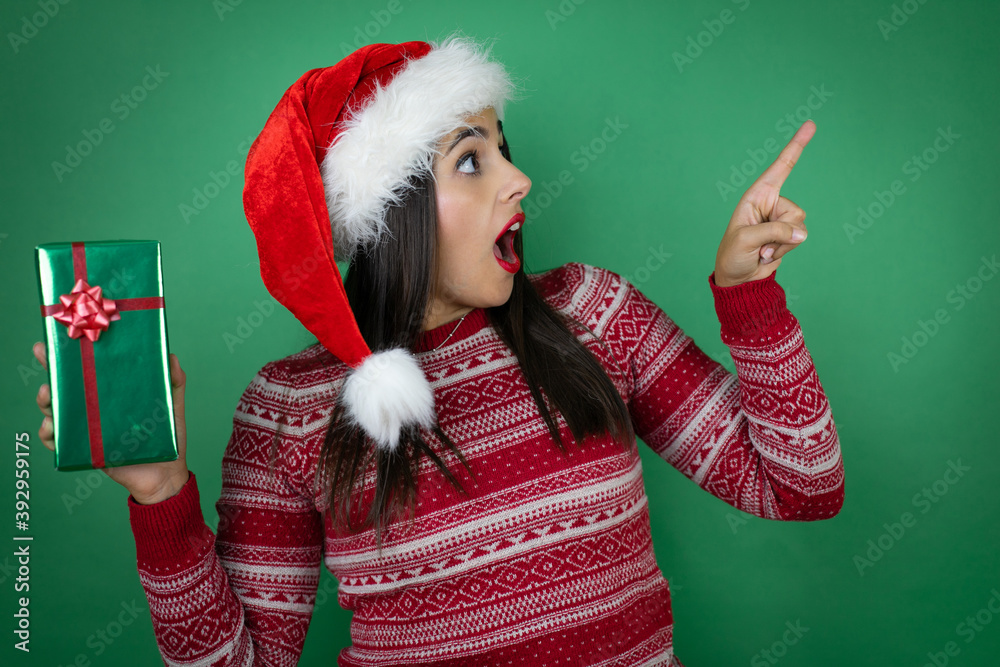 Young beautiful girl wearing Christmas Santa hat holding gift over isolated white background surprised and pointing side Young beautiful girl wearing Christmas Santa hat holding gift over isolated white background surprised and pointing side