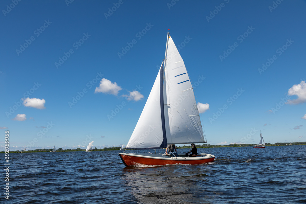 Fototapeta premium A red sailing boat on the Kagerplassen with 2 people sailing in the South-Holland municipality of Warmond in the Netherlands. On a beautiful day with a blue sky.
