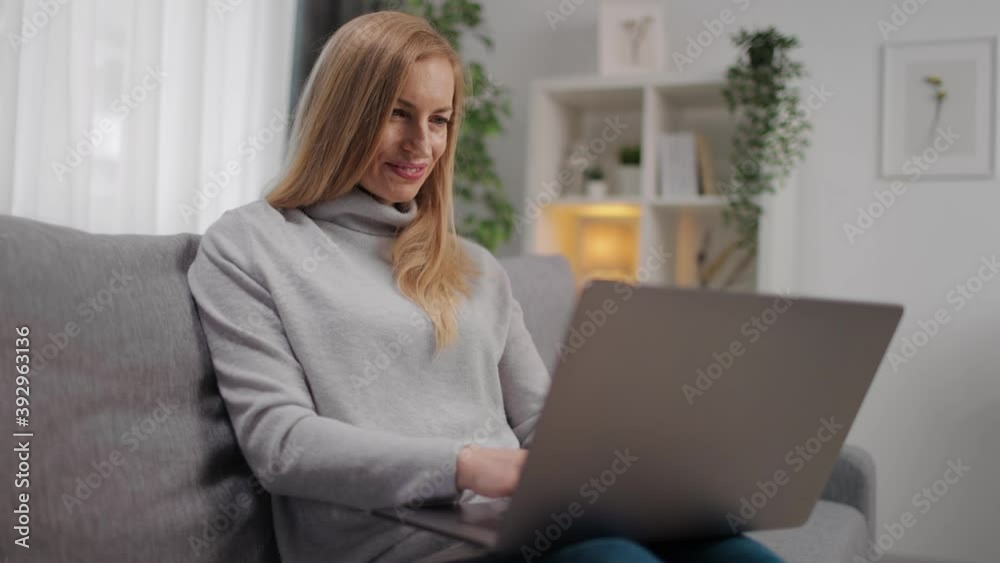 Beautiful female in casual outfit typing on wireless laptop while sitting on grey couch. Happy lady using modern gadget while staying at home. Beautiful female in casual outfit typing on wireless laptop while sitting on grey couch. Happy lady using modern gadget while staying at home.