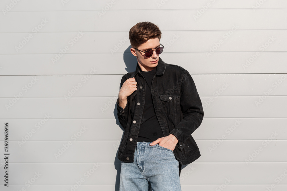 Stockfoto Fashion model handsome young man with trendy hairstyle in  sunglasses in black denim jacket in blue jeans with backpack posing near  vintage wall on street on sunny day. Attractive american guy