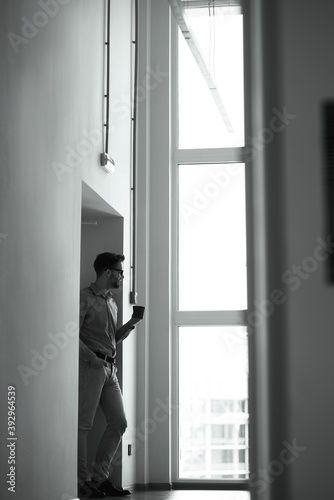 Vertical shot of a young businessman drinking fresh coffee while standing in the office and looking out the window, black black and white photo © Kostiantyn