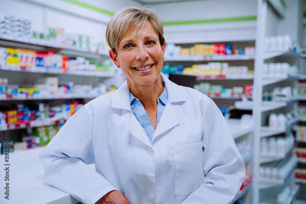 Smiling senior pharmacist looking happy leaning against medication counter standing in pharmacy 