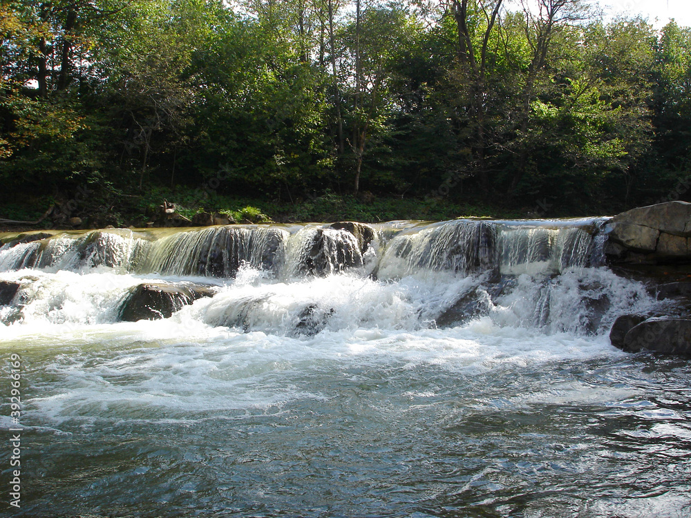 Fototapeta premium Stone threshold on a mountain river with streams of white water with foam and splashes among green trees