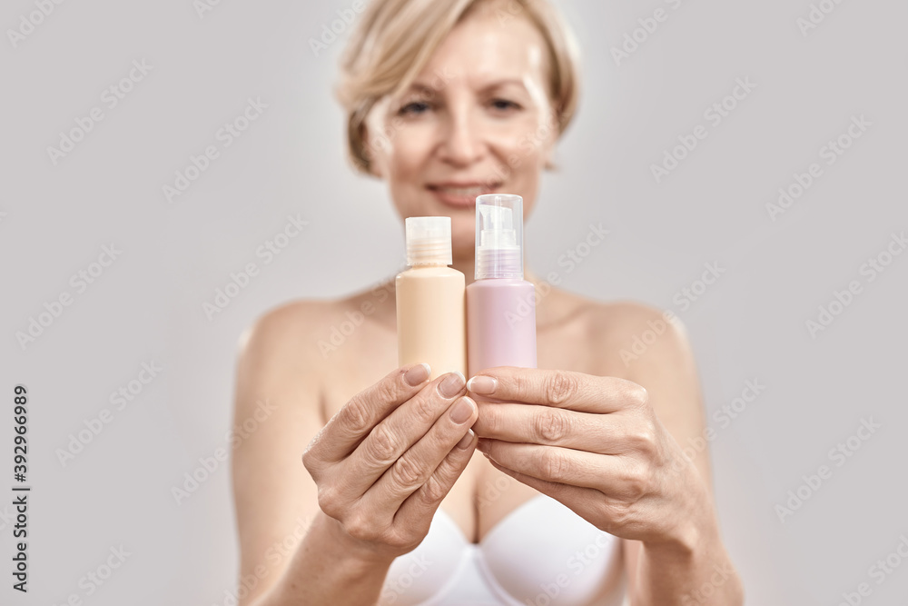 Close up of hands of middle aged woman holding two bottles of different cosmetic skincare products and comparing, choosing what to apply isolated over grey background Close up of hands of middle aged woman holding two bottles of different cosmetic skincare products and comparing, choosing what to apply isolated over grey background
