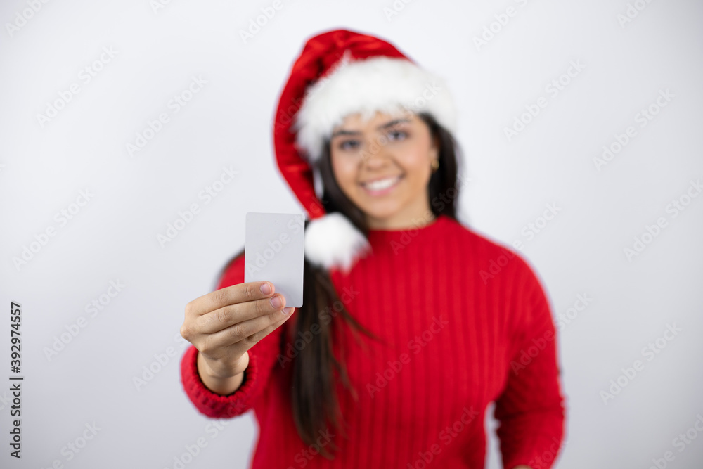 Young beautiful woman wearing a Santa hat over white background smiling and holding white card Young beautiful woman wearing a Santa hat over white background smiling and holding white card