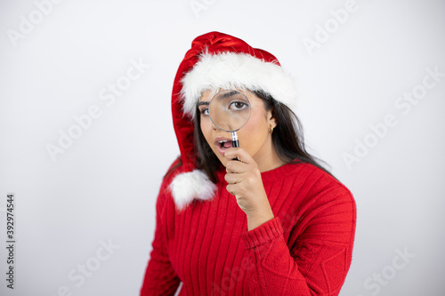 Young beautiful woman wearing a Santa hat over white background surprised looking through a magnifying glass