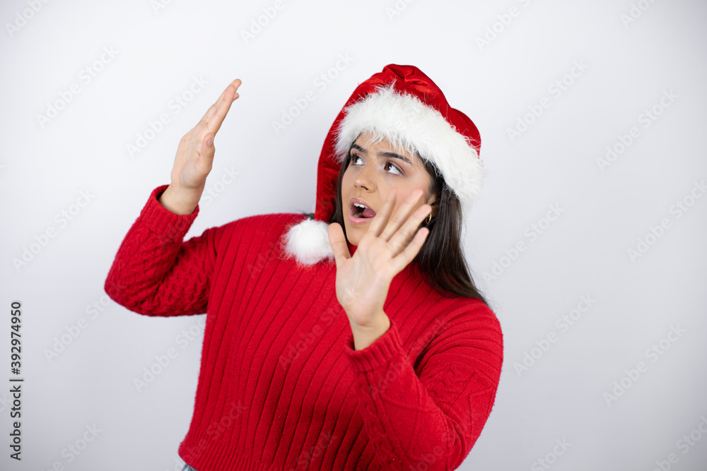 Young beautiful woman wearing a Santa hat over white background scared with her arms up like something falling from above Young beautiful woman wearing a Santa hat over white background scared with her arms up like something falling from above