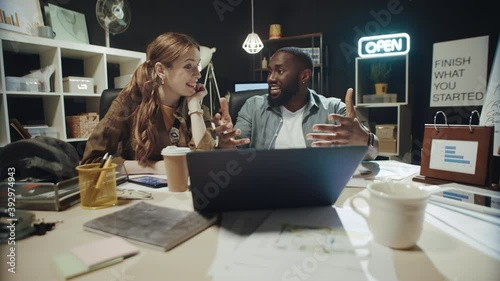 Smiling african american man telling story to businesswoman near laptop indoors.