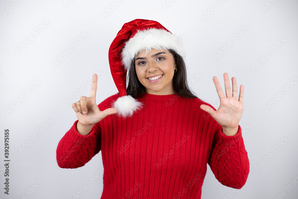 Young beautiful woman wearing a Santa hat over white background showing and pointing up with fingers number seven while smiling confident and happy Young beautiful woman wearing a Santa hat over white background showing and pointing up with fingers number seven while smiling confident and happy