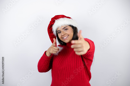 Young beautiful woman wearing a Santa hat over white background pointing to you and the camera with fingers  smiling positive and cheerful