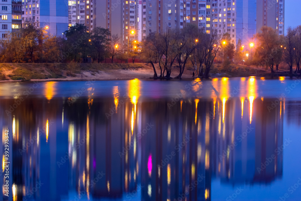 Silhouettes of multi-storey houses with bright light in the windows reflect on the blue water surface in the lake. Promenade by the lake at night