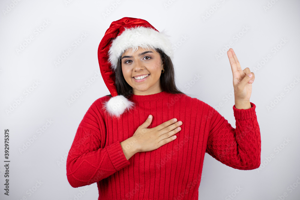Young beautiful woman wearing a Santa hat over white background smiling swearing with hand on chest and fingers up, making a loyalty promise oath Young beautiful woman wearing a Santa hat over white background smiling swearing with hand on chest and fingers up, making a loyalty promise oath