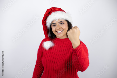 Young beautiful woman wearing a Santa hat over white background angry and mad raising fist frustrated and furious while shouting with anger. Rage and aggressive concept.