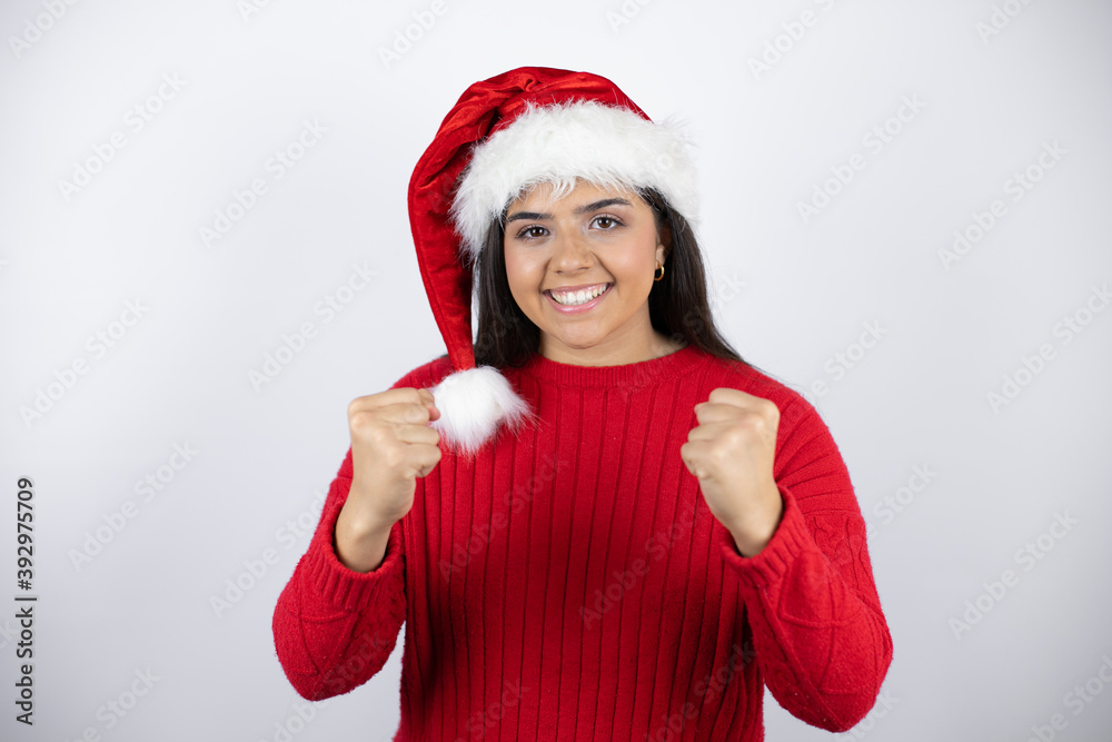 Young beautiful woman wearing a Santa hat over white background very happy and excited making winner gesture with raised arms, smiling and screaming for success. Young beautiful woman wearing a Santa hat over white background very happy and excited making winner gesture with raised arms, smiling and screaming for success.