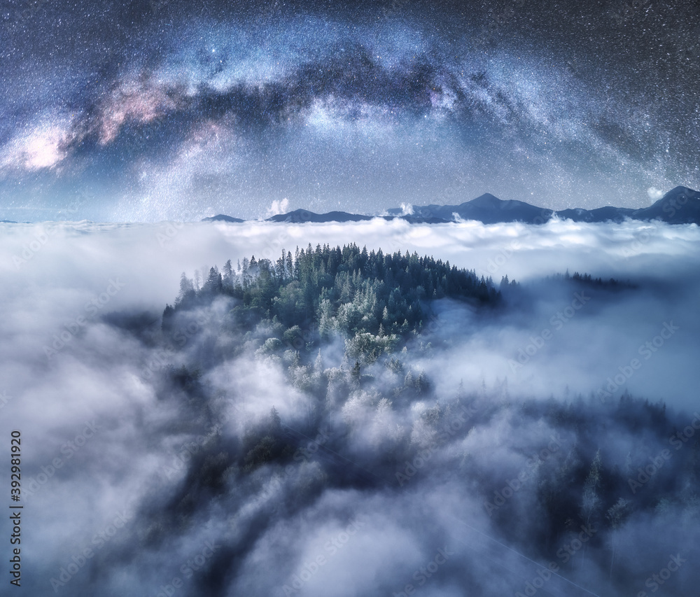 Milky Way arch over the mountains in low clouds at starry night in summer. Landscape with sky with stars, arched Milky Way, trees on the hill in fog, mountain peaks. Space and galaxy. Aerial view
