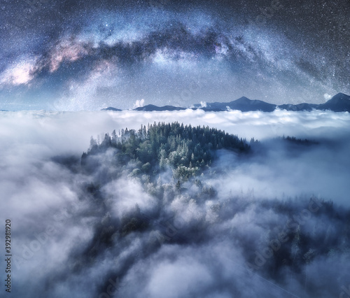 Milky Way arch over the mountains in low clouds at starry night in summer. Landscape with sky with stars, arched Milky Way, trees on the hill in fog, mountain peaks. Space and galaxy. Aerial view