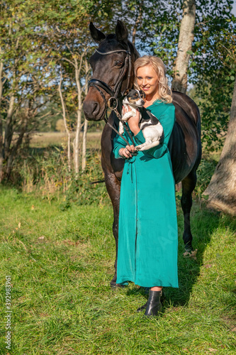 Beautiful blond smiling girl with her horse and dog in the forest. Wearing a green dress. Selective focus