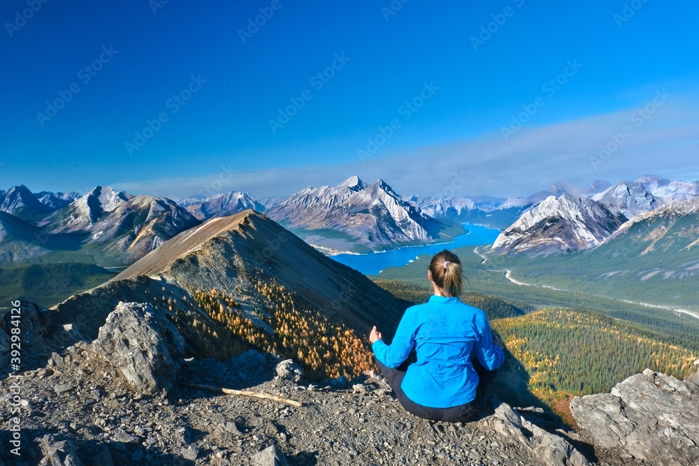 Naklejka premium Woman meditating on mountain top in front of beutiful lake and snowcapped peaks in Canadian Rockies. Alberta. Canada 