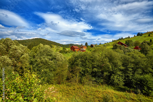 Summer landscape, rural houses on the slope of green mountains, against the background of a blue sky with clouds, trees and fir trees