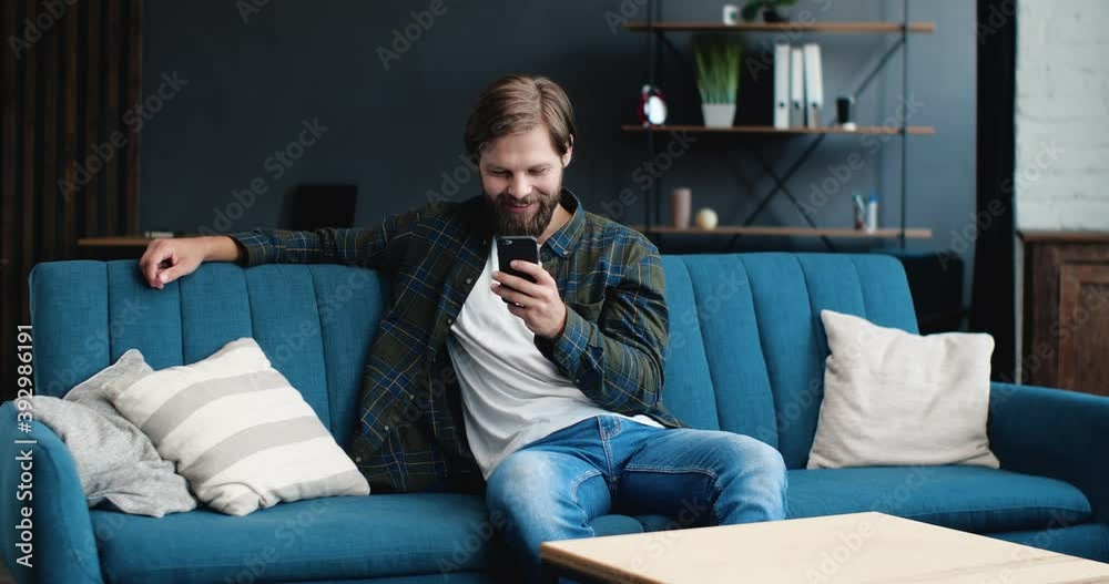 Portrait of Young Bearded Man is sitting at Home and chatting on his Smartphone, smiling Charmingly. Satisfied Man using Phone, texting Messages while relaxing on the Couch in Livingroom. Portrait of Young Bearded Man is sitting at Home and chatting on his Smartphone, smiling Charmingly. Satisfied Man using Phone, texting Messages while relaxing on the Couch in Livingroom.