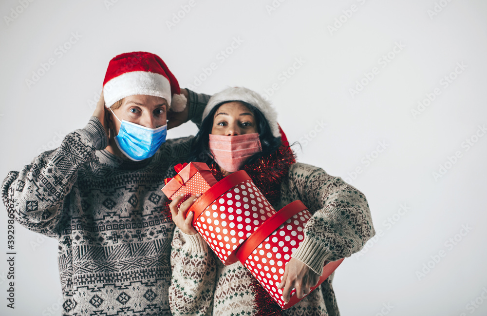 Young married couple with medical masks wearing santa hats with a gift in their hands wish each other a Merry Christmas