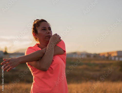An hispanic woman doing some stretch movements before do exercise in a park with the sunset behind her