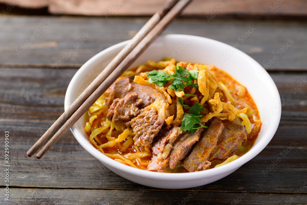 Northern Thai food (Khao Soi), Spicy curry noodles soup with beef in a bowl and chopsticks on wooden background Northern Thai food (Khao Soi), Spicy curry noodles soup with beef in a bowl and chopsticks on wooden background