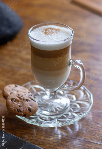 A coffee cappuccino cup and chocolate chips cookies on a wooden table with a book and a beret on it