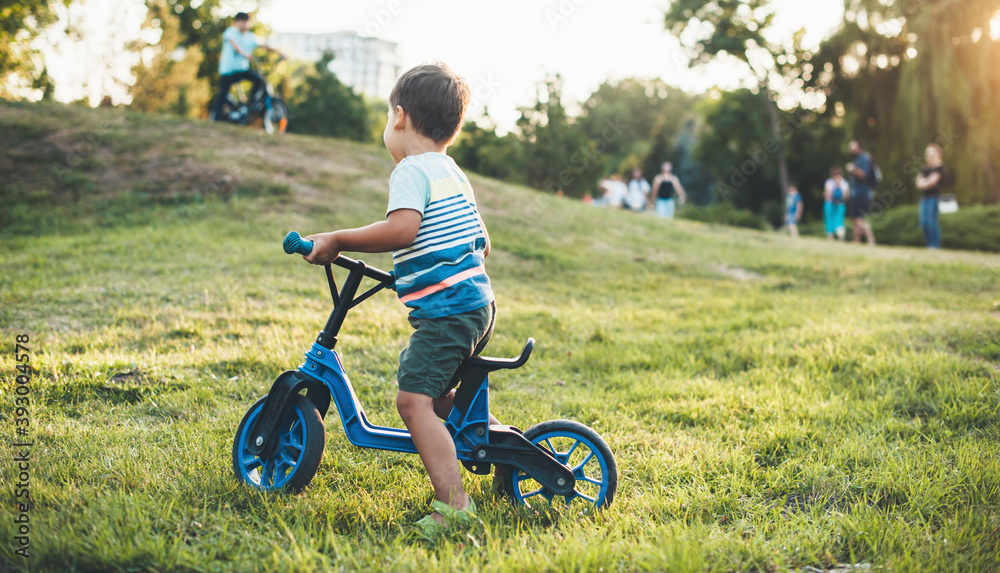 Small boy riding a bike in the park looking at other children walking with their parents Small boy riding a bike in the park looking at other children walking with their parents
