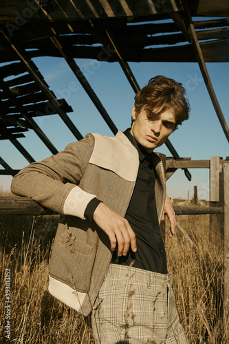 Portrait of young beautiful man in village area. Yellow field. Sun light, blue sky, autumn. Checkered pants, black shirt and jacket