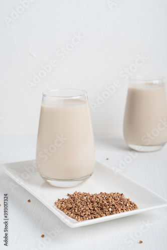 Buckwheat in form of seeds and glass of gluten-free milk which is source of protein, amino acids, manganese and vitamins for healthy dieting on rectangular plate on white wooden background. Vertical