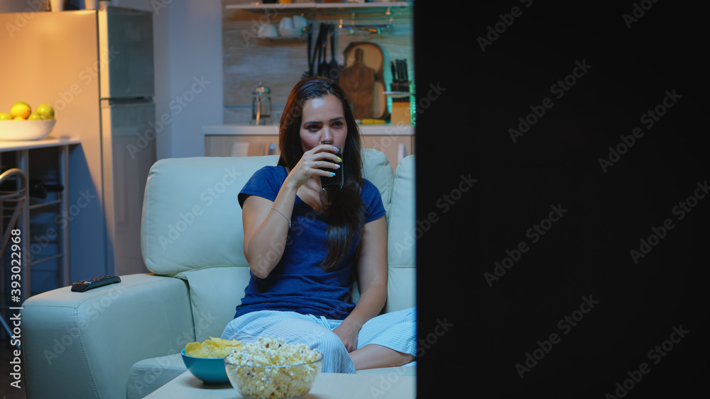 Naklejka premium Housewife relaxing at comedy show eating popcorn and drinking juice. Excited amused home alone woman enjoying the evening watching tv series at home sitting on comfortable couch dressed in pajamas.