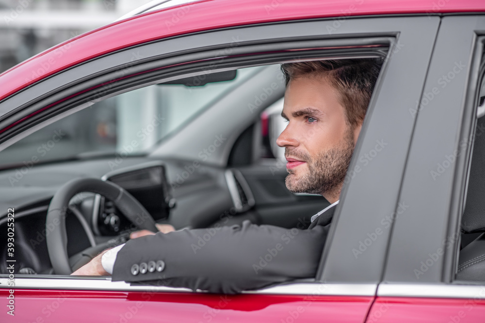Young bearded male sitting in red car, elbow out of window