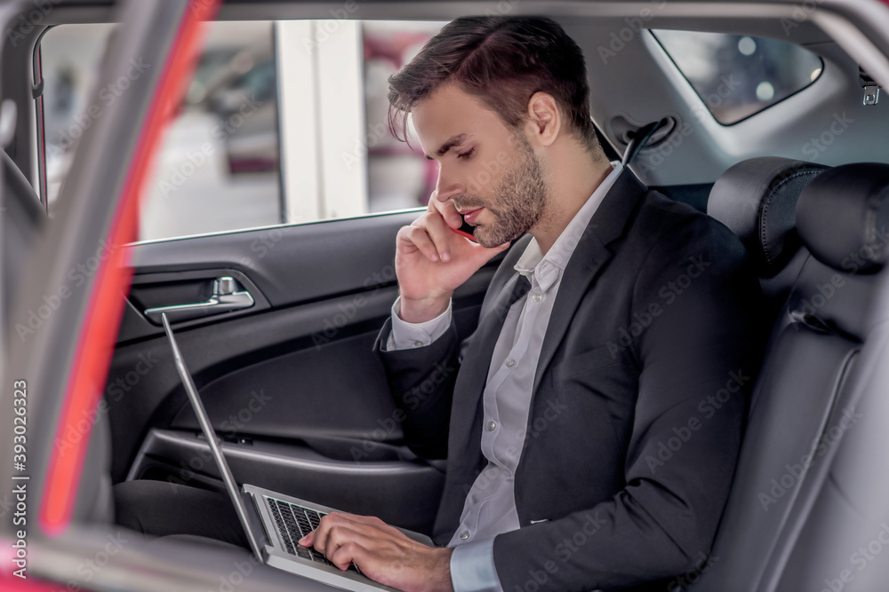 Brown-haired male sitting at backseat of car with laptop, talking on phone