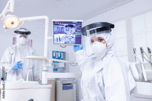 Patient pov at orthodontic doctor dicussing about teeth x-ray on digital screen.Stomatology specialist wearing protective suit against infection with coronavirus pointing at radiography. photo