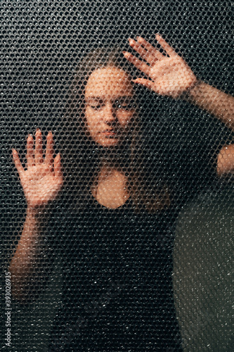 Pandemic self isolation. Quarantine loneliness. Covid-19 lockdown. Textured art portrait of depressed sad woman in black with closed eyes touching plastic bubble wrap wall in darkness.